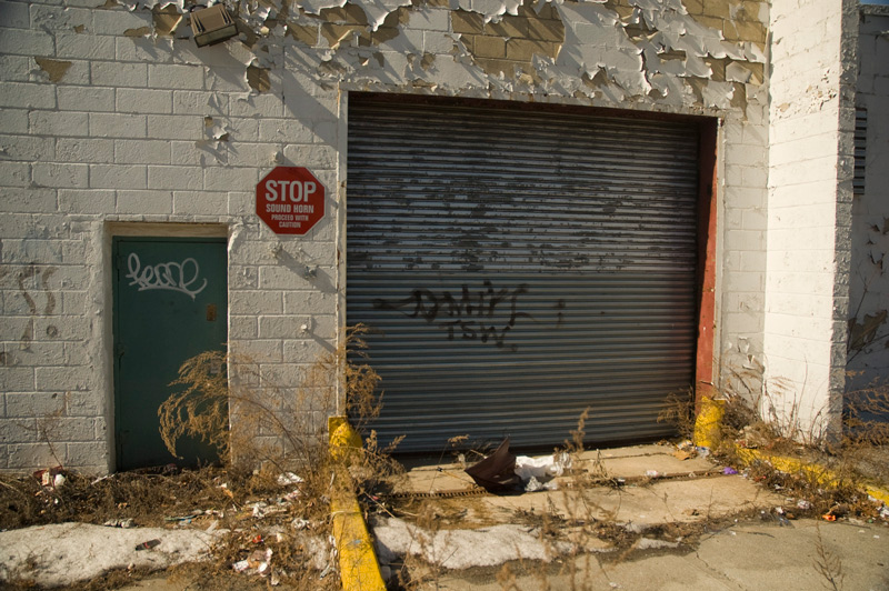 A warning sign on a cnider block building with peeling paint and weeds.