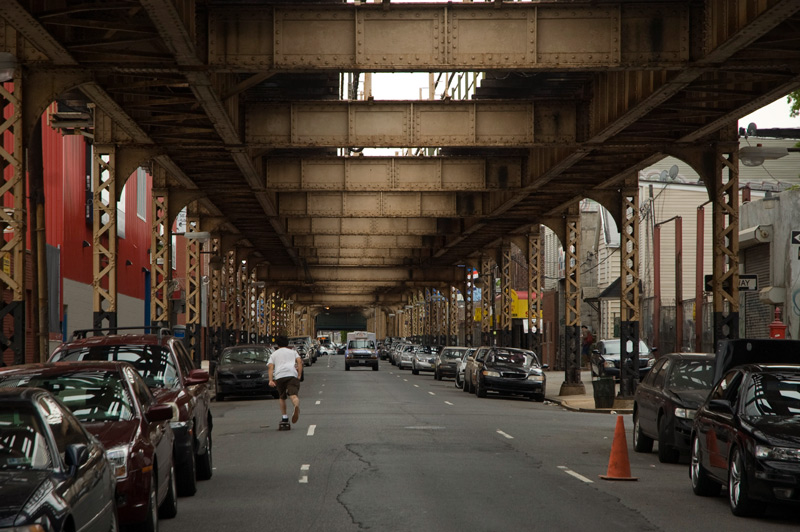 A man on a skateboard, under elevated subway tracks.