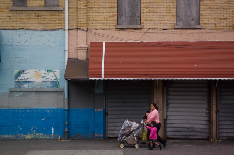A mother and four young children walk past a boarded up building.
