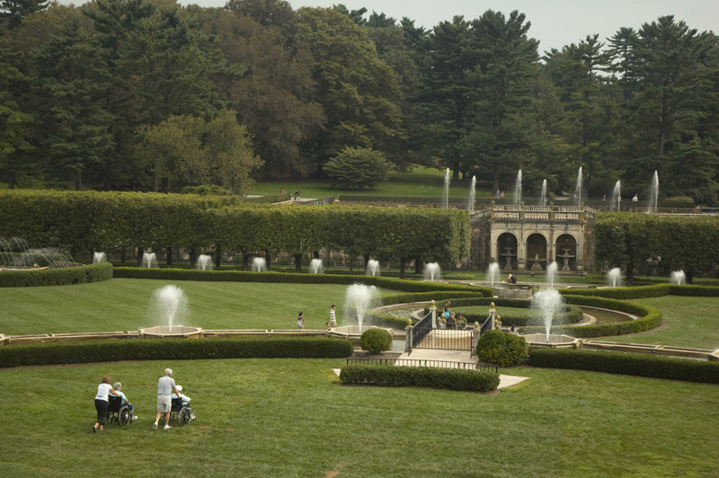 A pair of people push another pair in wheelchairs out to a trimmed garden with water fountains.