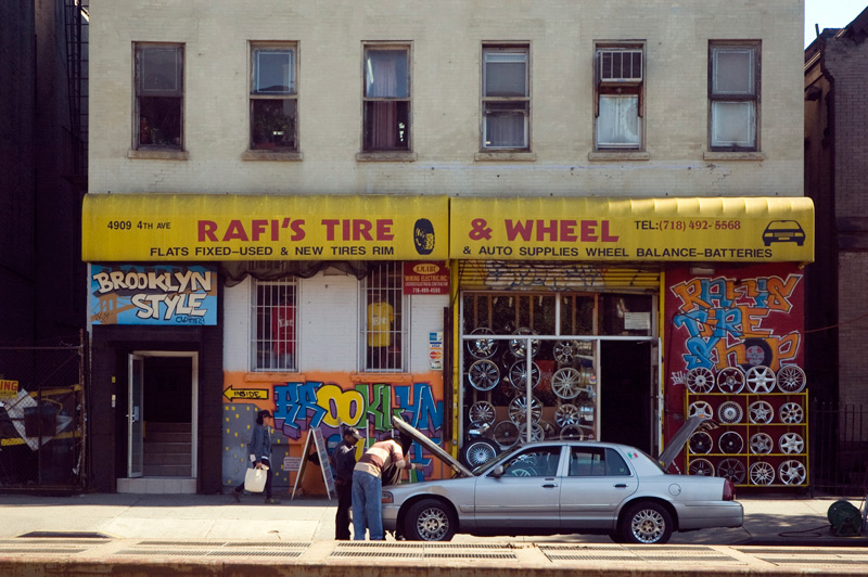 Two men huddle under a car hood in front of an auto supple store