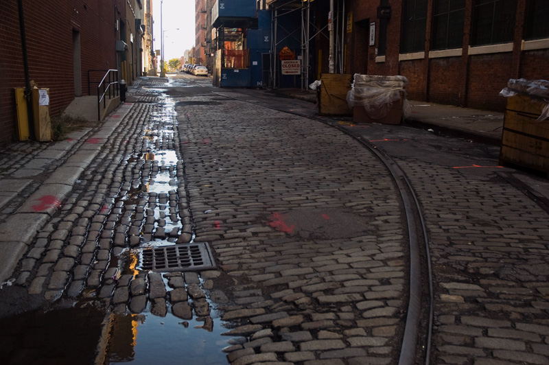 A street of paving stones, with railroad tracks and rivulets of water