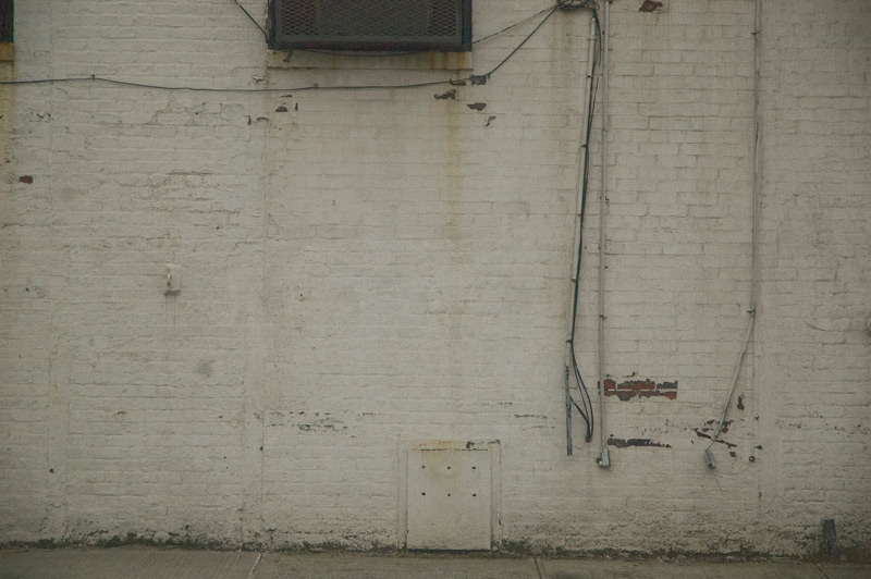 A white brick wall, with grates, metal plates, and wires.