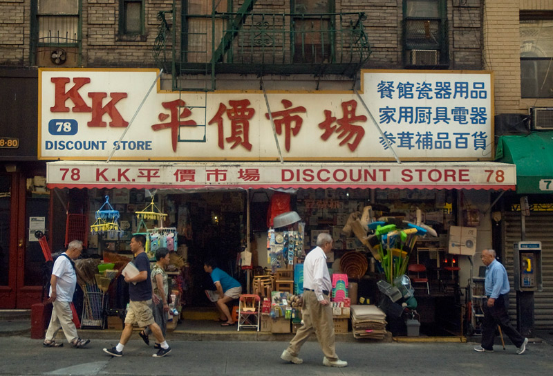 People passing a store selling brooms, mobiles, area rugs, odds and ends.