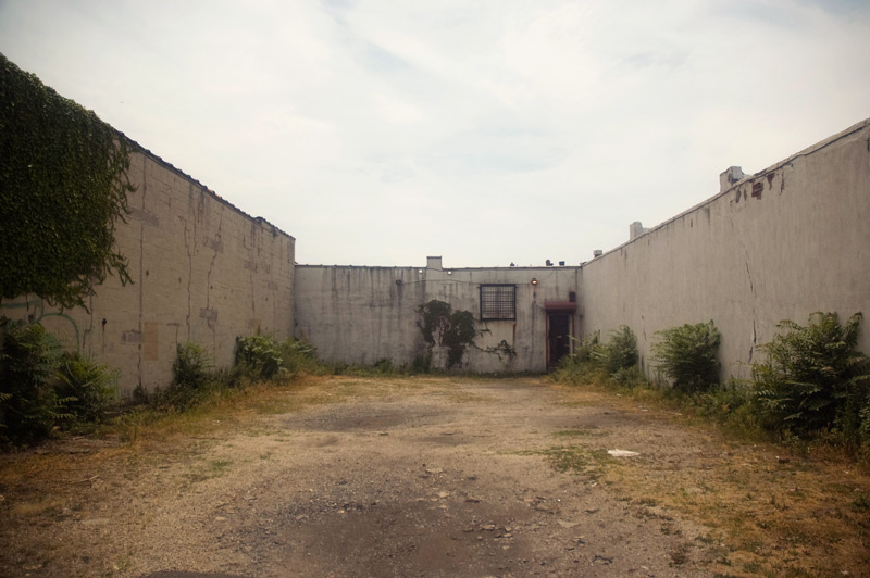 Three walls surround a yard with pavement and overgrown weeds.