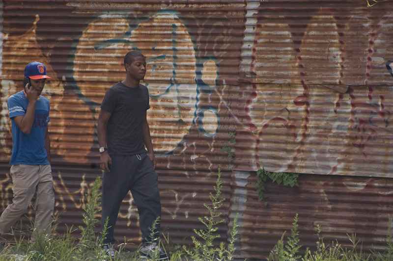 Two people walking past a corrugated metal fence.