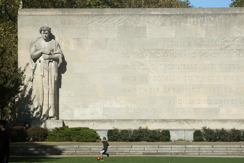 Child kicking a ball in front of a towering world war two memorial.