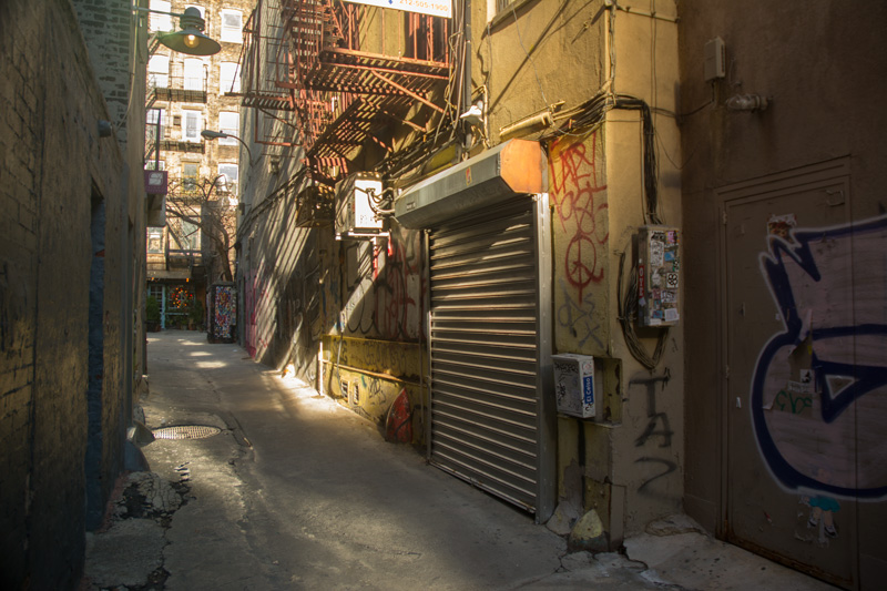 Sun shining into an alley, past rolling doors and fire escapes.