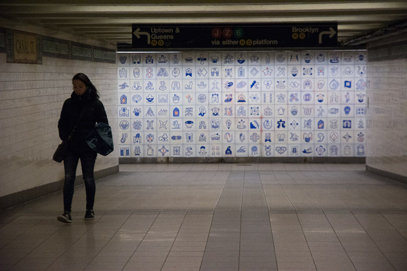 A woman walks up a passage, past a mural of tiles.