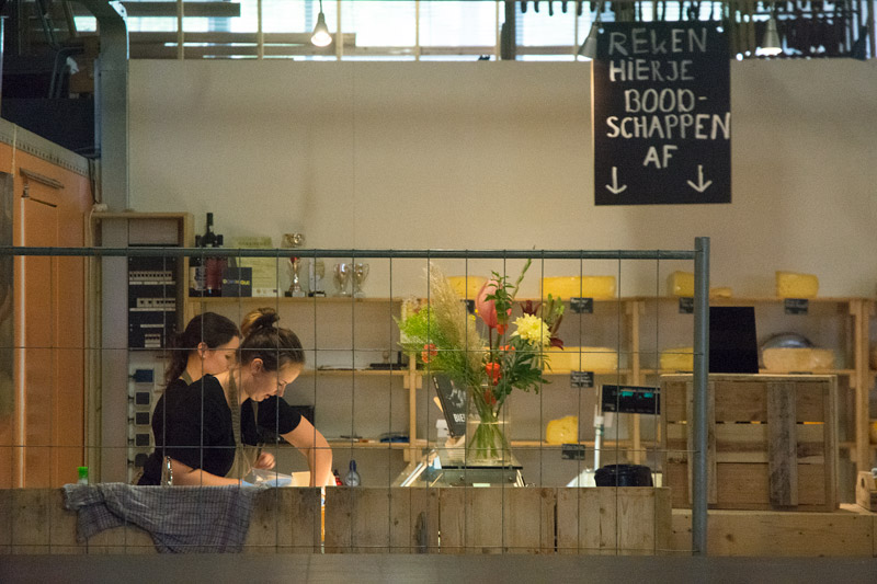 Two people behind the counter of a cheese shop, with wheels of cheese on shelves.