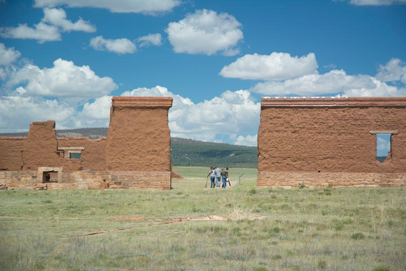 A group of people working in a gap between old adobe walls, amidst blue skies and prairies.