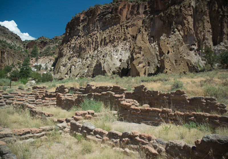 Ruins of a Native American village, below cliffs and a blue sky.