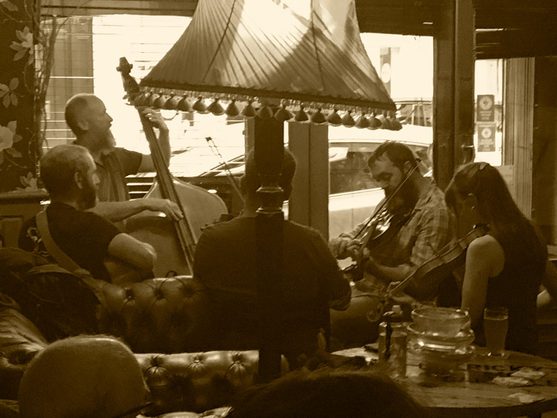 A quintet of 3 fiddles, a guitar, and an upright bass in a bar with padded seats.