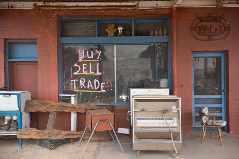 Beat-up goods in front of a junk store, whose hand-painted sign says 'BUY! SELL! TRADE!'.