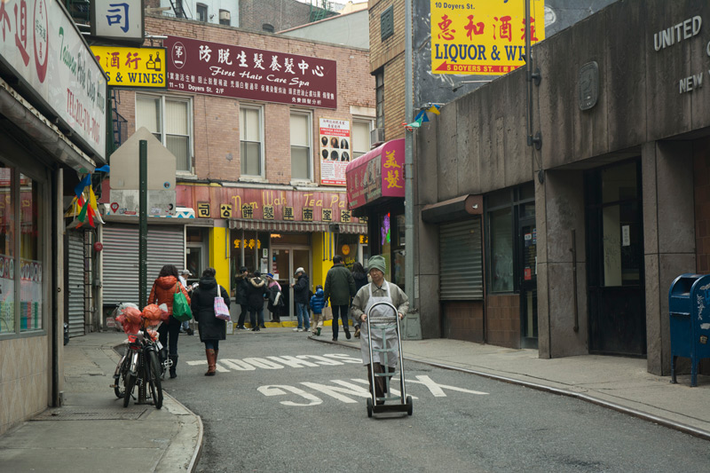 A man in an apron is pushing an empty hand truck down the middle of the street.