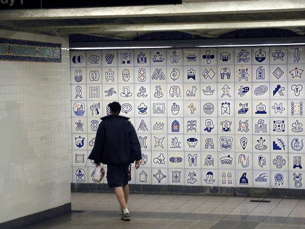 A man goes down
a hall, with a wall of blue and white tiles beyond him.