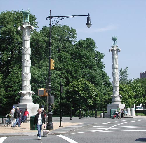 Two tall, ornate columns on opposite sides of a
road, with huge green trees and a woman walking her
dog.