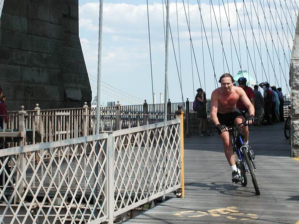 A shirtless man on a bicycle careens around
a corner on the wooden platform of the Brooklyn
Bridge.