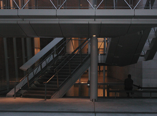 A man sits on a bench at dusk, as lights start to
go on in the lobby beyond him.