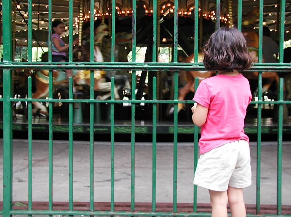 A little girl stands outside a fence, looking in
at a carousel.