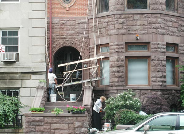 A painter raises his scaffold on a brownstone,
while the owner gardens out front.