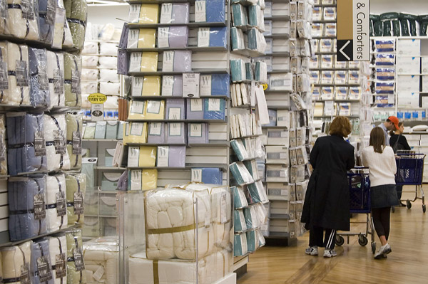 Two people roll their shopping cart past stacks of linens.