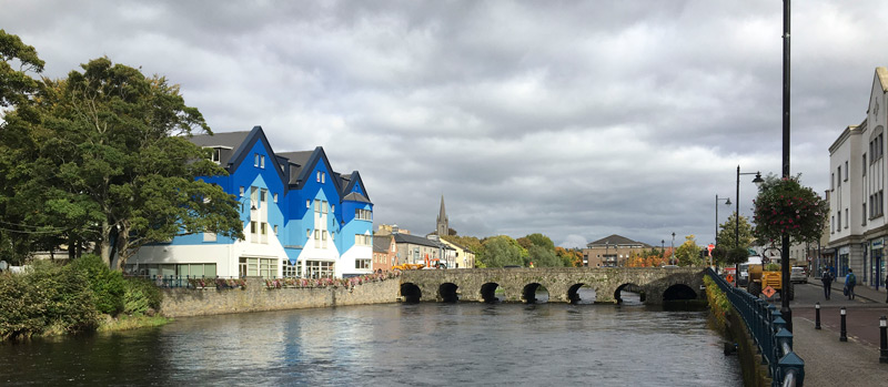 Three blue and white buildings sit next to a stone bridge and the Garavogue River.